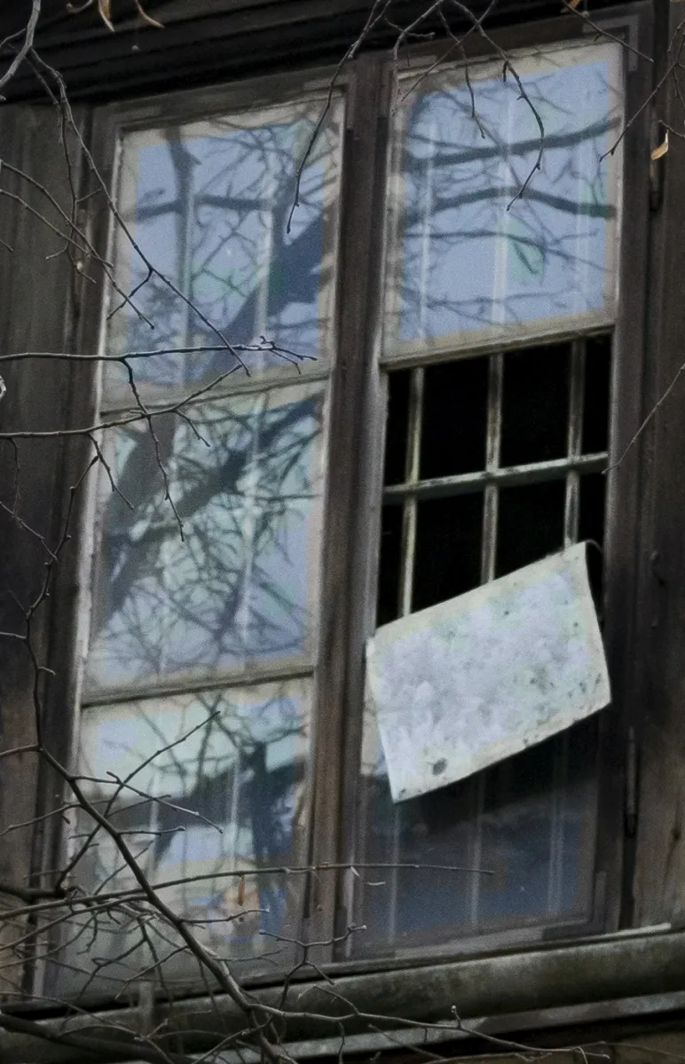 Weathered multi-pane window with one broken section and a board hanging from it